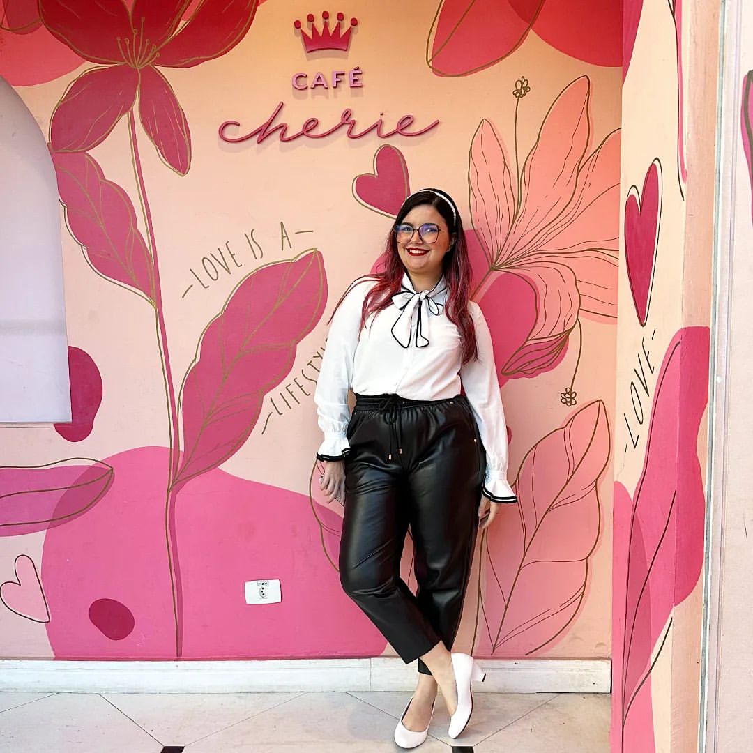 Posando em frente à fachada do Café Cherie, onde há sua logo de coroa no alto, que é toda rosa com desenhos de folhas na mesma cor. Na foto estou usando um arco branco nos cabelos rosa, um batom vermelho, óculos de grau, camisa branca com um laço na gola, calça preta que imita couro e sapato de salto branco.