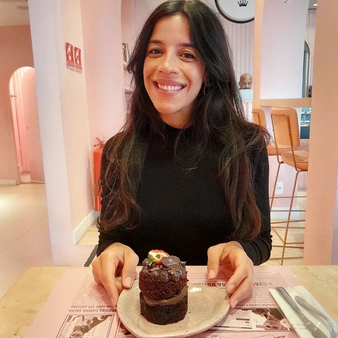 Foto de uma mulher de cabelos longos e escuros e blusa preta, sentada à mesa, sorrindo. À sua frente há um mini naked cake de chocolate, com cobertura e recheio do mesmo sabor.