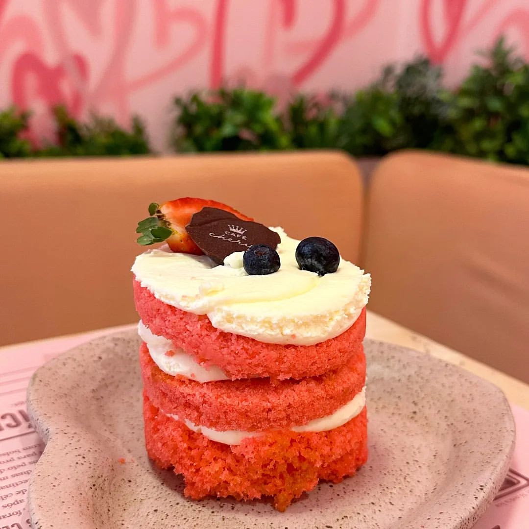No mesmo ambiente anterior, com a parede de corações ao fundo, é possível ver uma parte da superfície da mesa onde há um prato em forma de coração contendo um mini naked cake cor de rosa com recheio e cobertura brancos. Em cima da cobertura há alguns mirtilos e um morango.