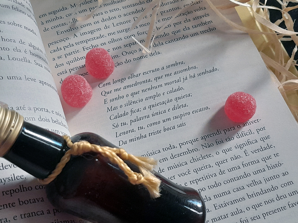 Livro aberto em página onde há um trecho de O Corvo, de Edgar Allan Poe, onde cita a personagem Lenore Dove. As jujubas e garrafa estão por cima do mesmo, em volta da citação.