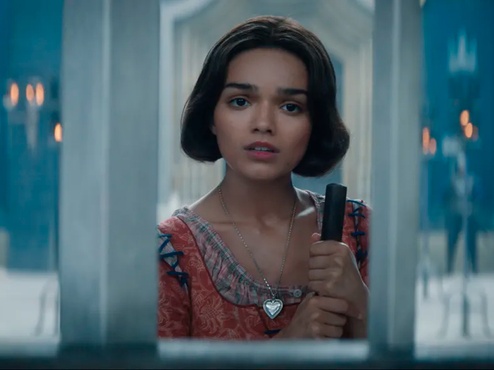 Cena do filme Branca de Neve onde a atriz Rachel Zegler aparece como a personagem vestindo roupa vermelha de criada, cabelos escuros curtos e expressão triste. Ela segura uma vassoura e está atrás de grades, podendo ser visto do busto para cima.