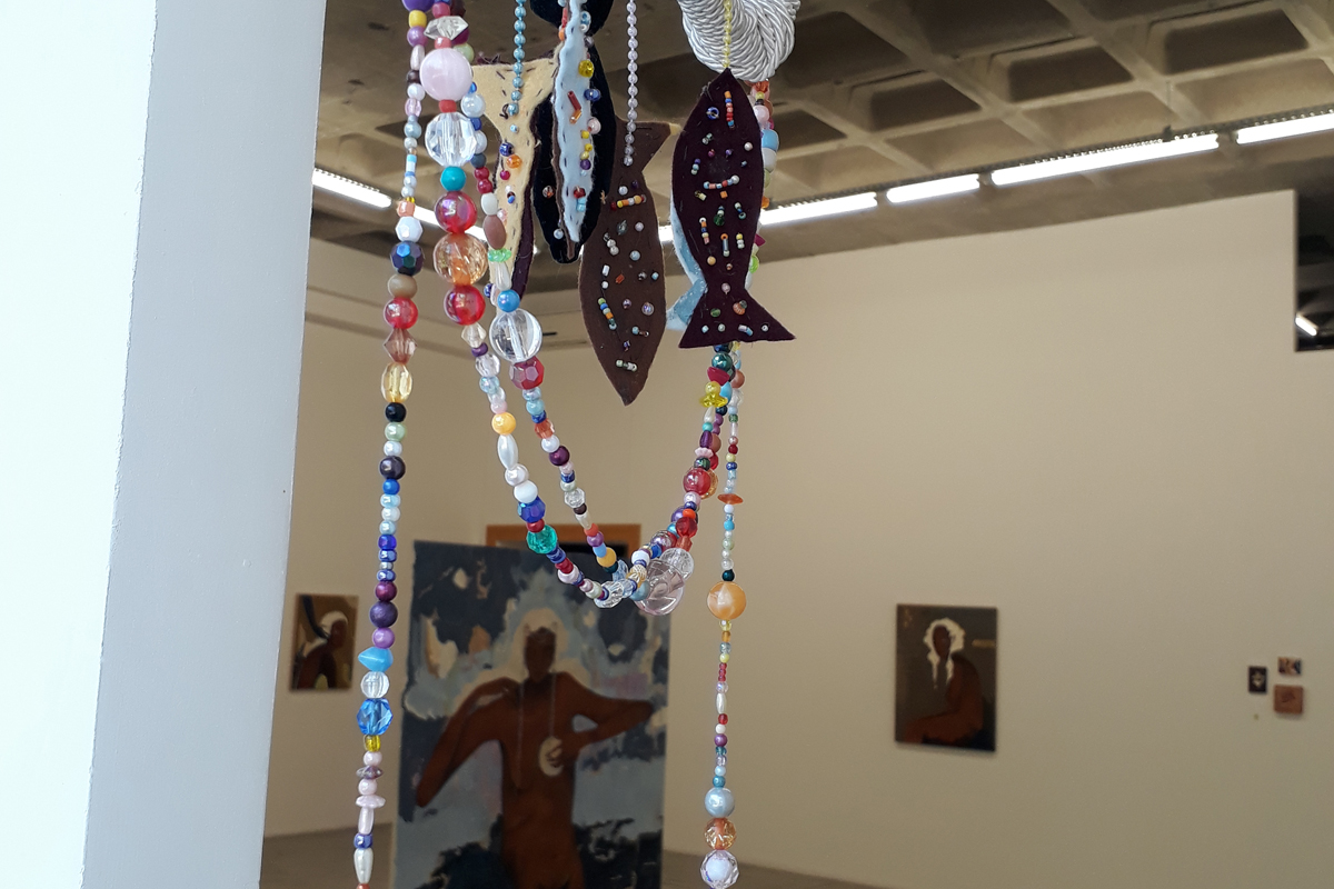 Móbile feito com miçangas e tecidos em forma de peixe em primeiro plano, com a sala principal da exposição ao fundo.