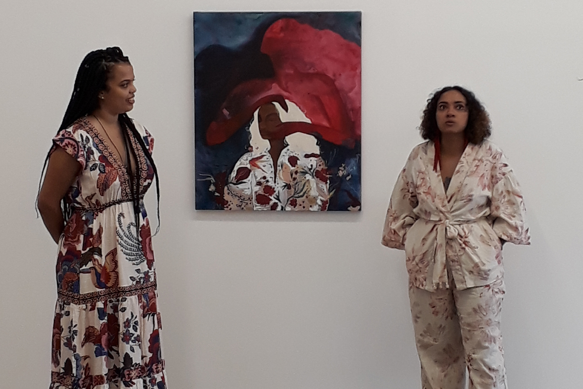 Duas mulheres negras dando uma palestra com uma pintura ao centro, sendo elas Lorraine Mendes, curadora,, e Jess Vieira, artista