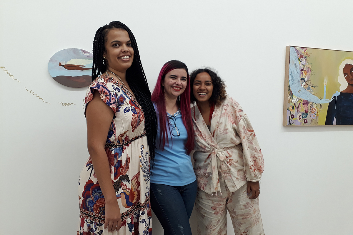 Três mulheres posando diante das obras, duas de pele negra e cabelos escuros e uma de pele clara e cabelos rosa, que está ao centro. Elas sorriem para a câmera, abraçadas. Elas são Lorraine Mendes, Luly Lage e Jess Vieira