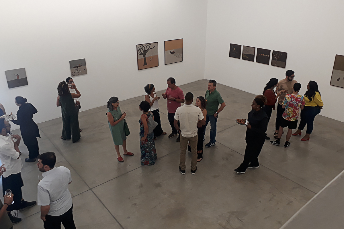 Vista do andar de cima do salão expositivo durante a abertura, onde várias pessoas conversam, olham as obras nas paredes e são servidas por garçonetes que vestem preto.