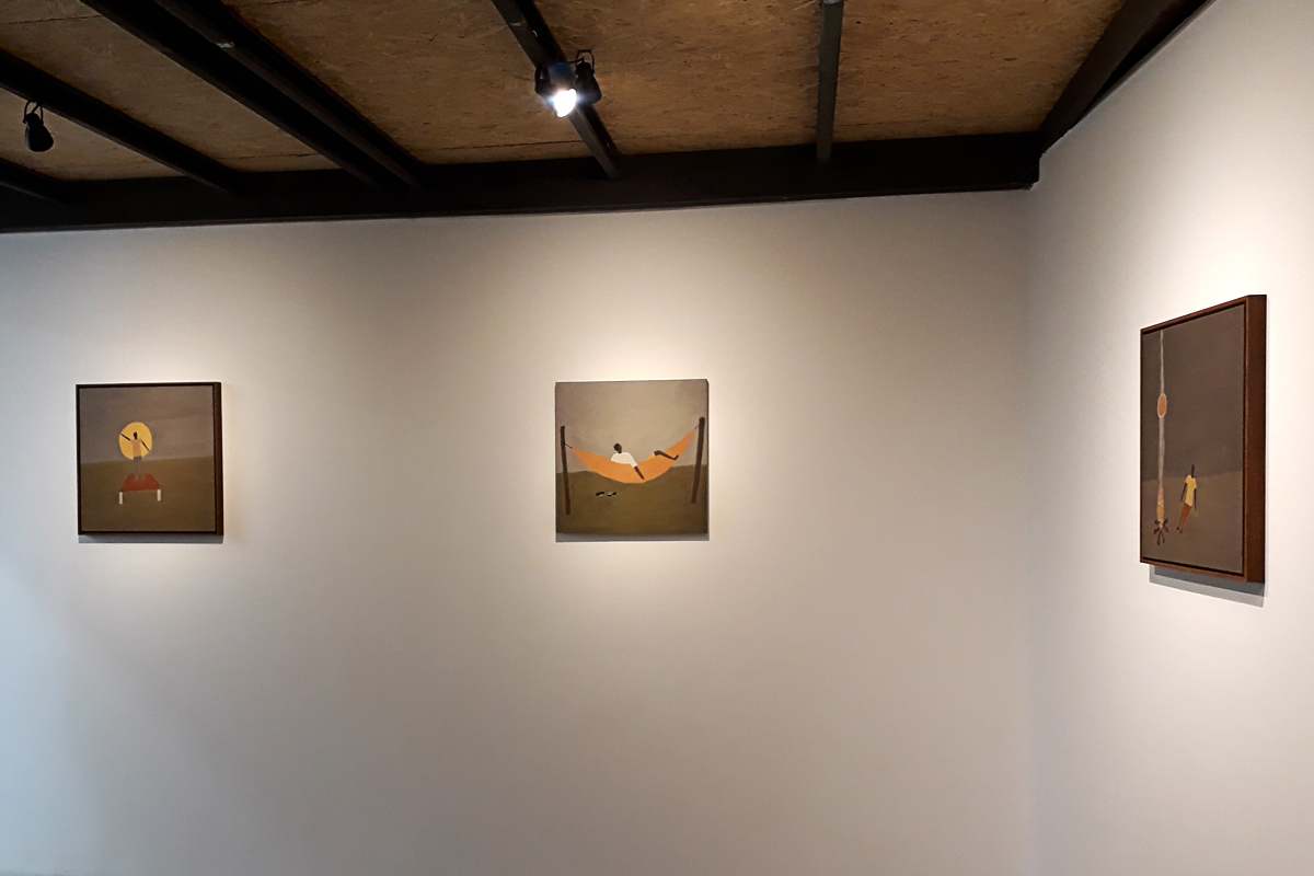 Segundo espaço expositivo, de iluminação menos clara, com três pinturas na parede, a primeira de uma pessoa pintando um Sol, a segunda de um homem na rede e a terceira, que aparece quase de lado, de um homem sentado diante de uma grande fogueira acesa.