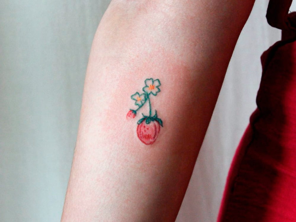 A primeira das tatuagens é na parte interna do praço, abaixo da dobra do cotovelo, representando um galho com dois morangos, um maior e outro bem menor, em tons de rosa e vermelho simulando giz de cera. Os cabos são vermelhos e terminam em flores brancas de miolo amarelo.