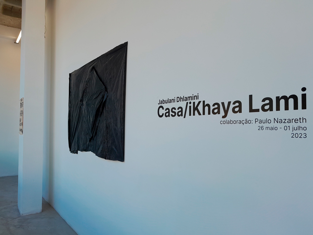 Fachada da exposição com título, nome do artista e duas obras de Paulo Nazareth, uma bandeira do Brasil toda preta feita de lona e uma placa indicando a conexão de Belo Horizonte e Joanesburgo.