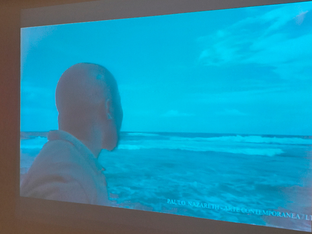 Vídeo de Paulo Nazareth onde se vê Jabulani, em preto e branco, de costas para a câmera e de frente ao mar.