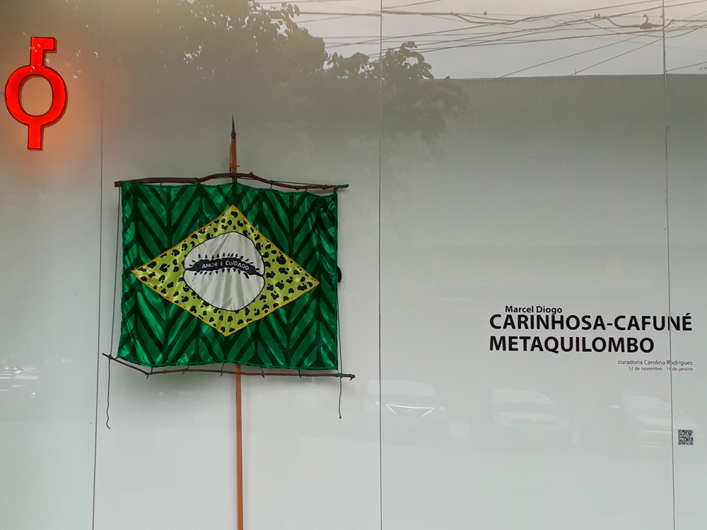 Fachada da exposição onde há uma obra de arte representando uma nova versão da bandeira do Brasil onde há estampa nas cores verde e amarelo, o branco no lugar do azul e os dizerem AMOR E CUIDADO na faixa. Ao seu lado, o nome de Marcel Diogo, o título Carinhosa-Cafuné MetaQuilombo e a Curadoria de Carolina Rodrigues.