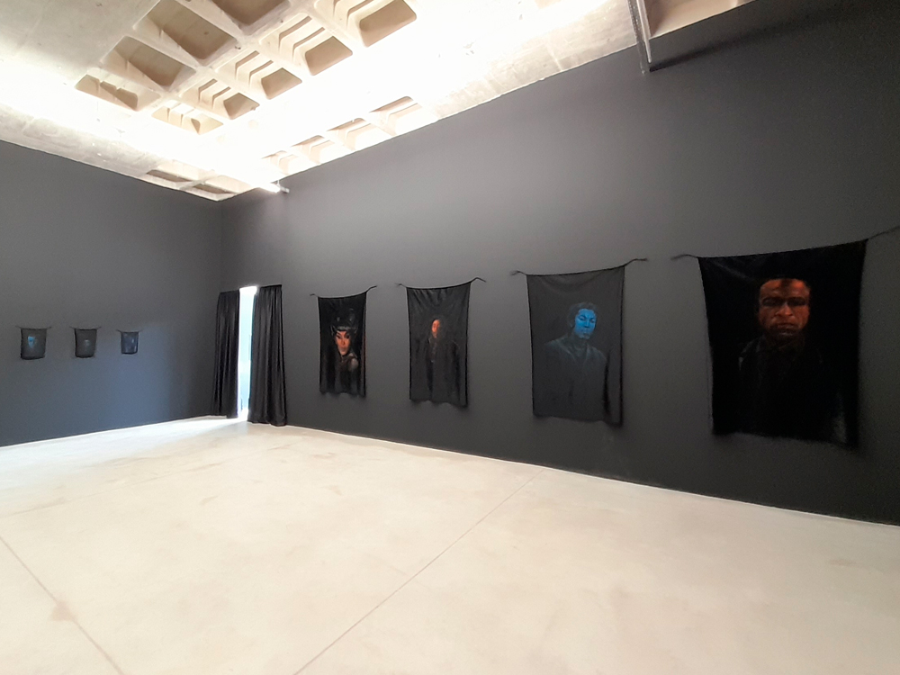 Salão expositivo contando pinturas da série carinhosa-cafuné de Marcel Diogo, a parede do fundo tem três pinturas de pequeno porte e, na outra, quatro pinturas médias representando Elza Soares, Bob Marley, Milton do Nascimento (que é a única de tinta azul, enquanto as outras têm tinta alaranjada) e Frantz Fallon. A pelúcia de todas as obras é preta, assim como a parede da galeria.