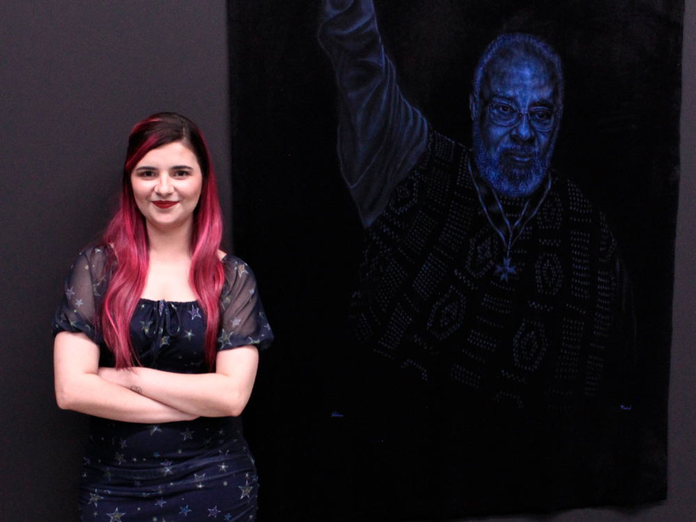 Foto de Luly Lage de braços cruzados, encostada em uma parede preta ao lado de uma pintura em pelúcia de Abdias do Nascimento com o braço direito erguido.
