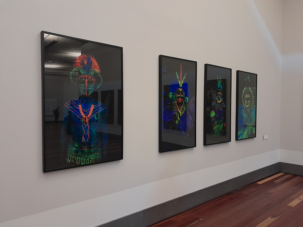 Parede com quatro fotografias com fundo escuro, preto, e rostos de pessoas também escuros, onde se destaca pinturas em tons de neon.