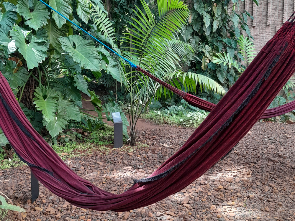 Instalação com redes vermelhas na porta da galeria, com piso coberto de pedras e paisagismo de plantas ao fundo