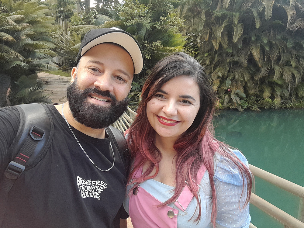 Um homem e uma mulher abraçados, ele de camisa, boné e barba pretos, ela de cabelos e jardineira cor de rosa e blusa azul, os dois sorrindo e posando para a câmera, com o paisagismo de Inhotim visto ao fundo