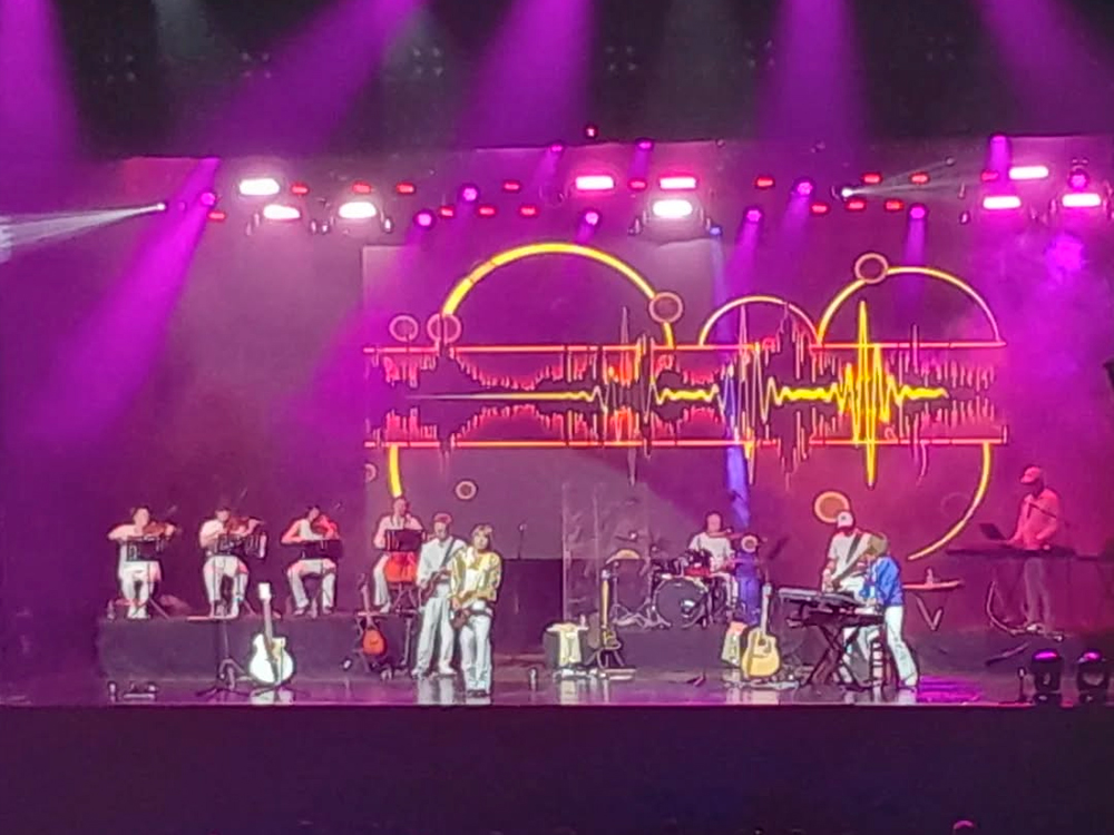Show ao vivo com músicos no palco, todos vestidos de branco com detalhes coloridos. Ao fundo, um grande telão exibe uma imagem gráfica de batimentos cardíacos estilizados em vermelho, amarelo e laranja, sobre linhas e círculos interligados. A iluminação é predominantemente roxa. Instrumentos como teclado, guitarras e violinos estão visíveis, com integrantes tocando ou em pé.