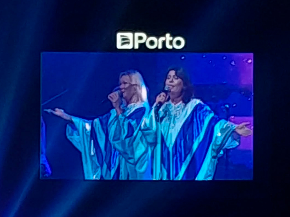 Telão exibe duas cantoras no palco vestidas com túnicas azuis brilhantes em estilo retrô, com detalhes claros. Elas estão de braços abertos, cantando com microfones em mãos, iluminadas por luz azulada. Acima do telão, vê-se a palavra “Porto”, indicando o nome do patrocinador do evento. O fundo é escuro, com luzes suaves e atmosfera de show.
