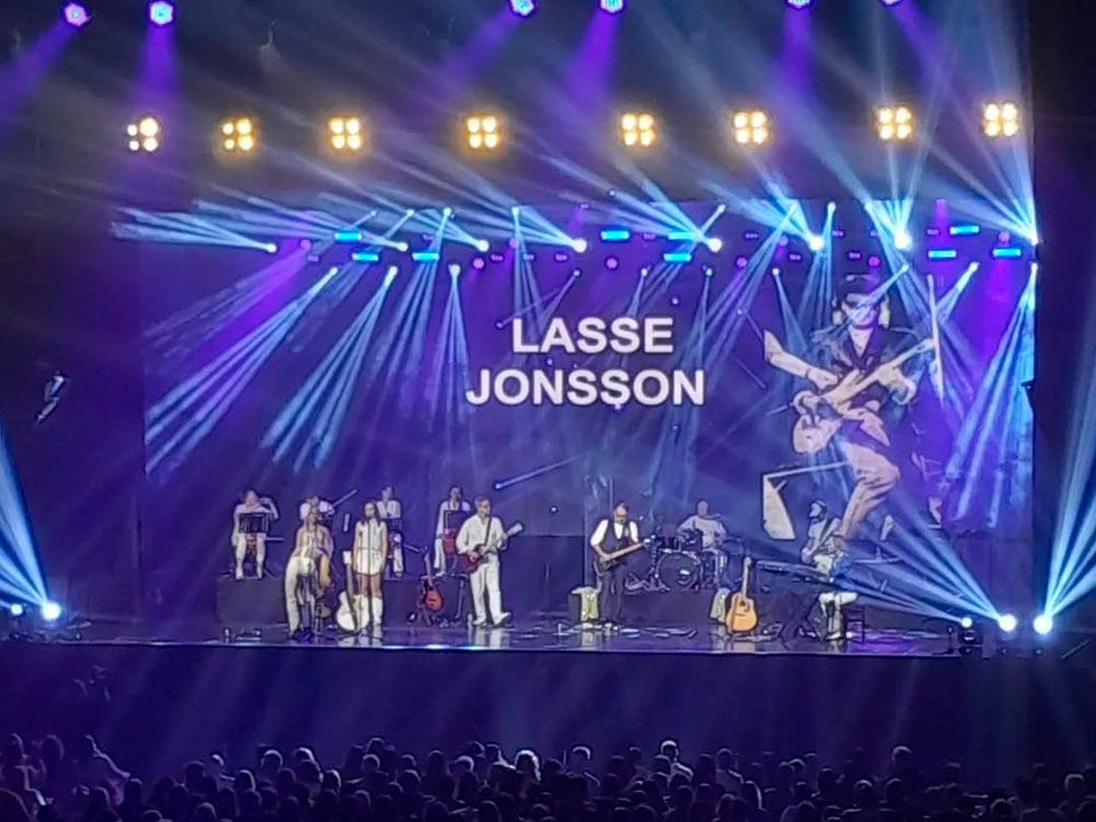 Palco iluminado com luzes roxas e brancas em feixes direcionados ao público. No centro, aparece o nome “Lasse Jonsson” em letras brancas sobre um fundo escuro, com uma imagem estilizada de um guitarrista em tons de roxo e bege à direita. A banda está posicionada ao fundo, com integrantes distribuídos em diferentes instrumentos — cordas, teclado, bateria e sopros. Várias pessoas no palco estão vestidas de branco, criando um visual harmônico e retrô. A plateia aparece em silhueta na parte inferior da imagem.