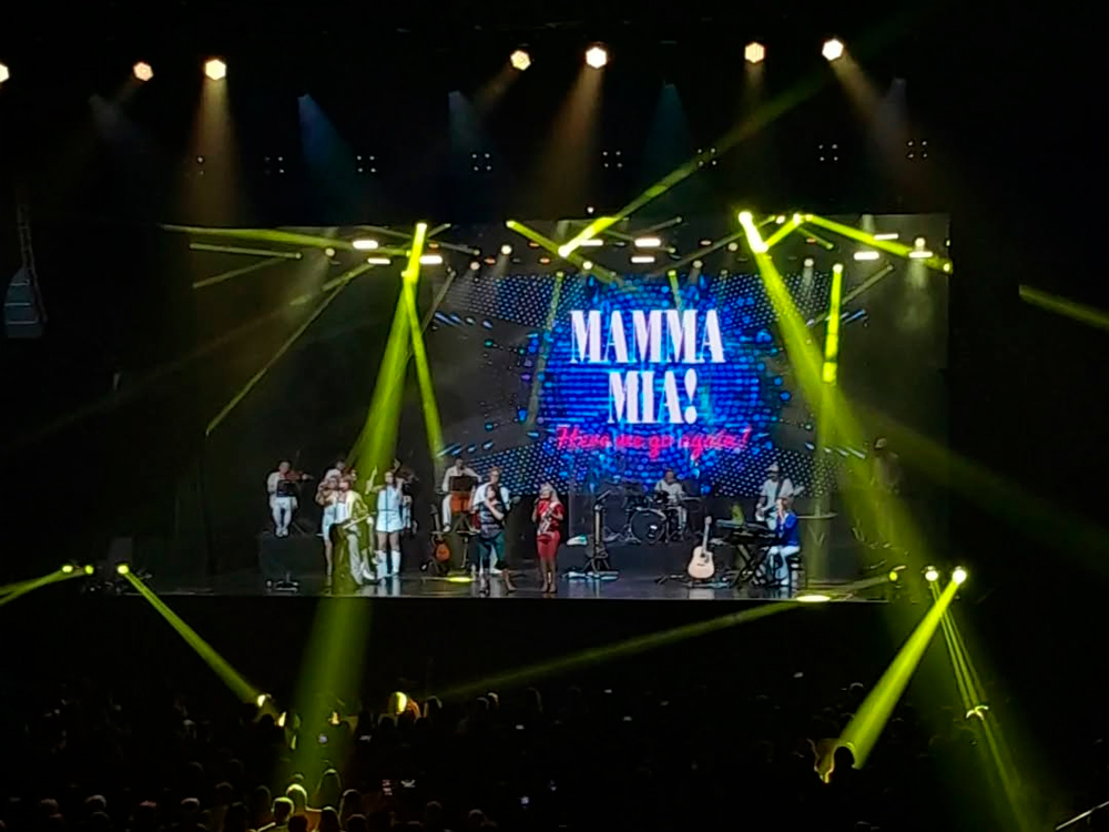 Palco iluminado com luzes amarelas vibrantes e um telão ao fundo exibindo a frase “MAMMA MIA! Here we go again!” em letras grandes e coloridas sobre um fundo azul pontilhado. No centro do palco, duas cantoras em figurinos inspirados nos anos 1970 — uma de vermelho e outra de preto com brilho — dividem os vocais. Ao redor delas, músicos e backing vocals completam a formação da banda tributo ao ABBA, com instrumentos como guitarra, teclado, violino e bateria. A plateia aparece em silhueta, com alguns braços erguidos e celulares levantados. A atmosfera é festiva e nostálgica.