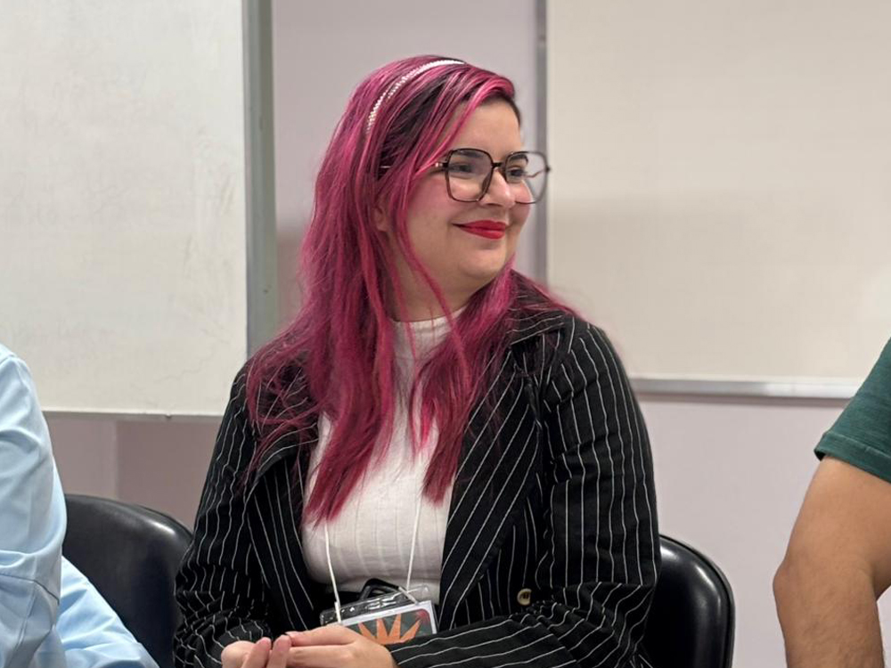 Sentada diante de um quadro branco, vestida com uma blusa branca e blazer preto com riscas brancas, um arco branco nos cabelos e batom vermelho. Na foto estou sorrindo, olhando para uma pessoa ao meu lado, o corpo virado de frente pra câmera com o rosto em perfil por causa disso.