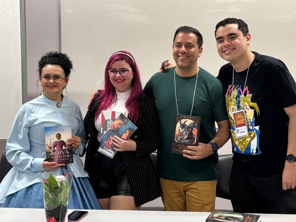 Os quatro de pé, atrás da mesa, posando para a foto, cada um com alguns livros nas mãos, na mesma ordem da foto anterior. Todos estão sorrindo.