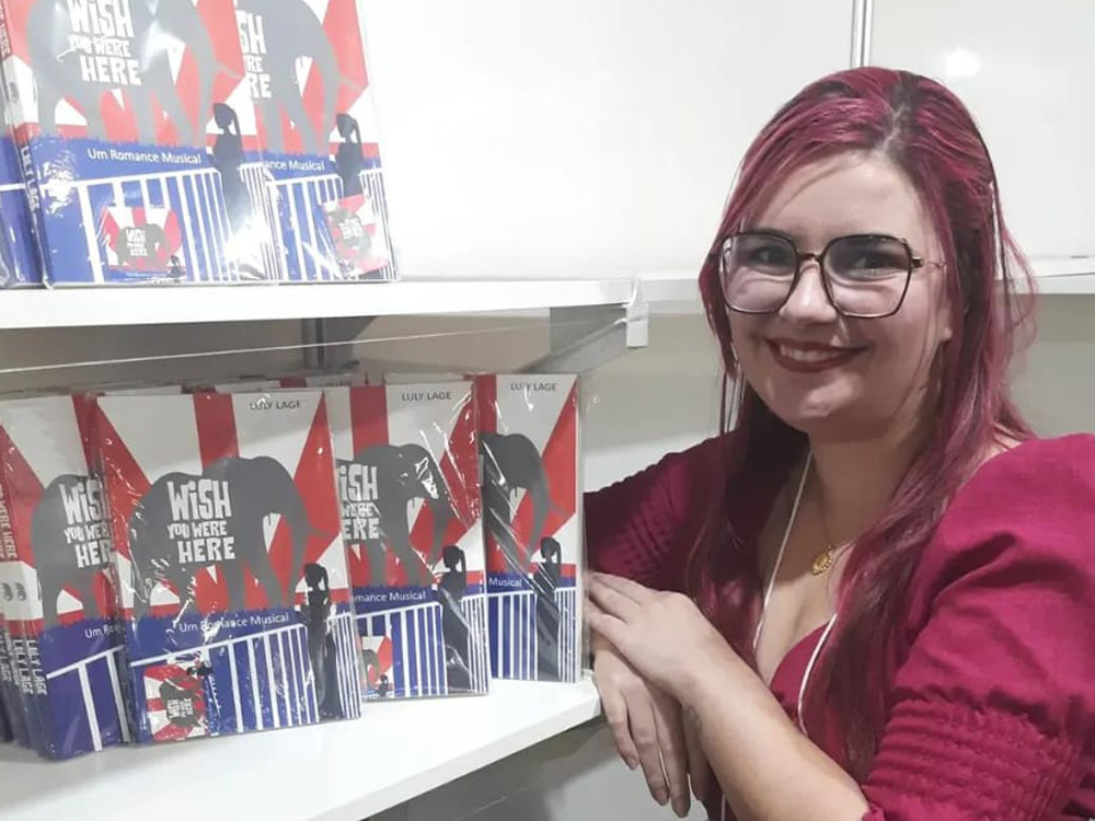Com a mesma roupa da foto destaque do post, de pé apoiada na estande onde estão os exemplares do livro Wish You Were Here, sorrindo para a câmera.