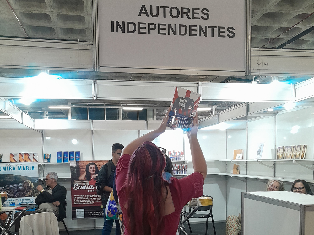 De costas, levantando o livro Wish You Were Here perto da testeira onde está sinalizado o estande de Autores Independentes da Bienal. Ao fundo, os outros autores interagem, alguns sentados e outros de pé.
