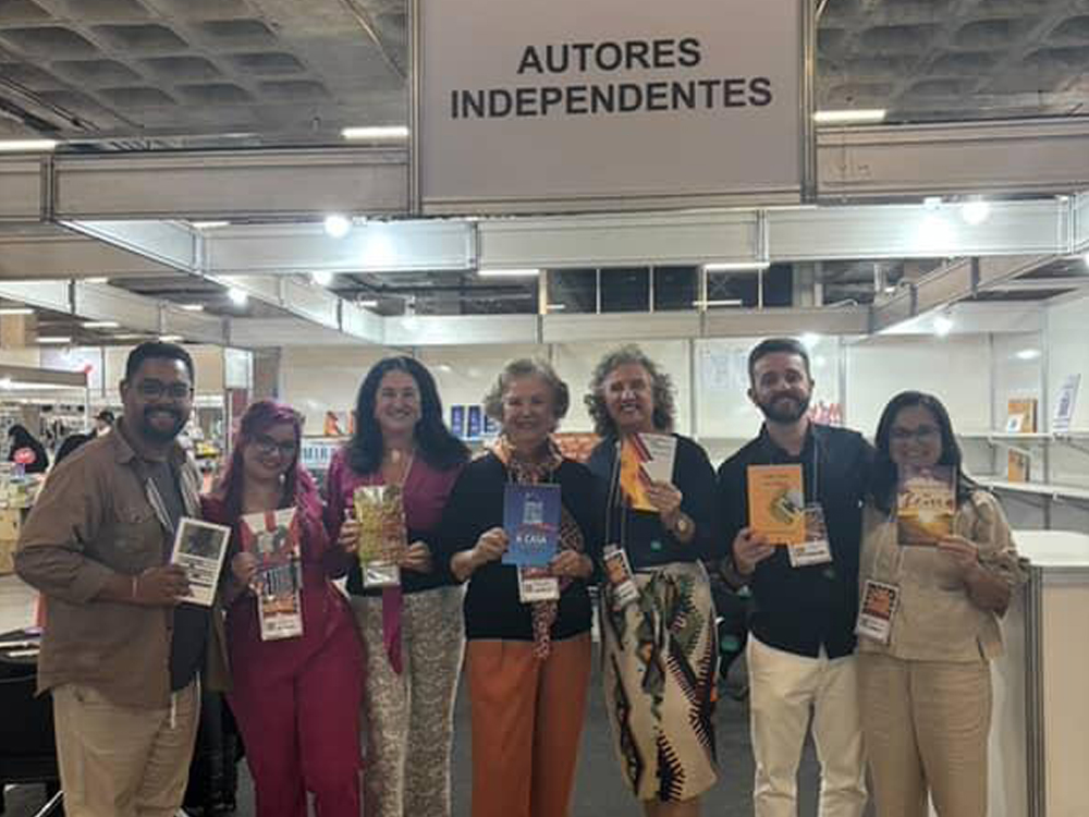 Todos os autores participantes de pé, lado a lado, em frente ao estande, segurando seus livros e portando seus crachás da Bienal.
