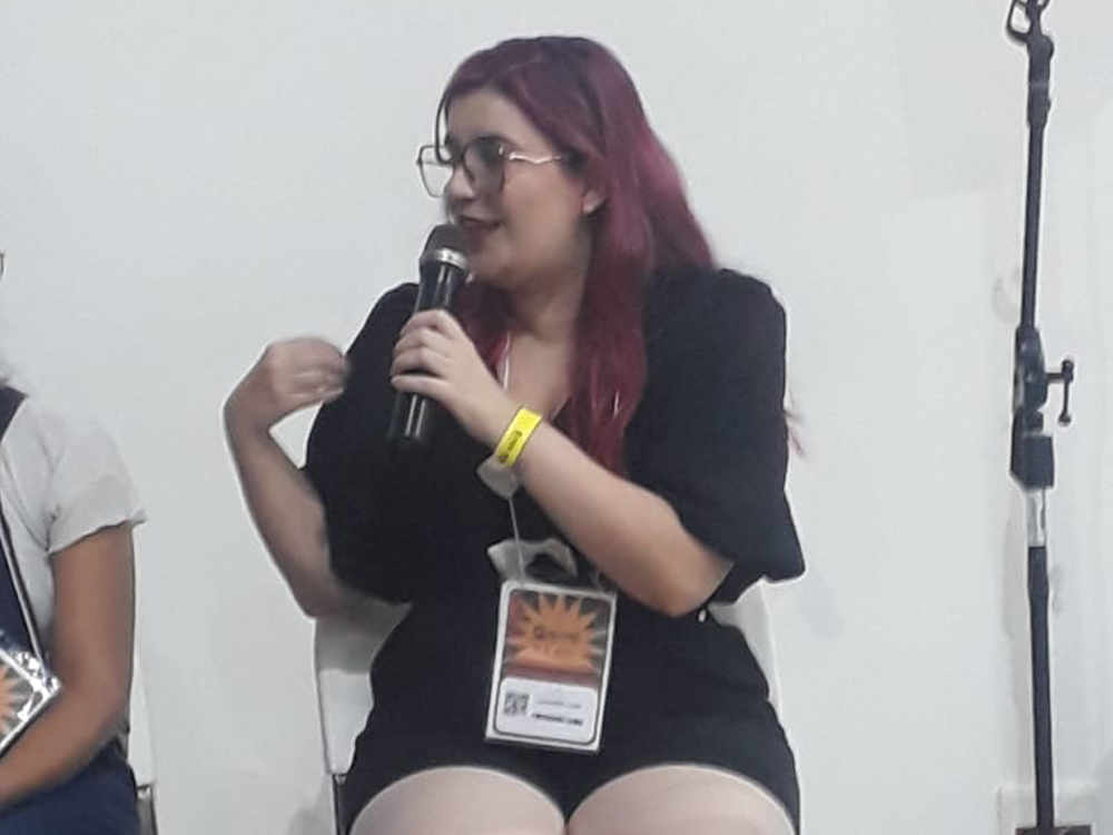 Sentada com o microfone na mão, vestindo um macaquinho preto com três laços brancos na frente, uma meia calça branca, um arco preto e o crachá da Bienal.
