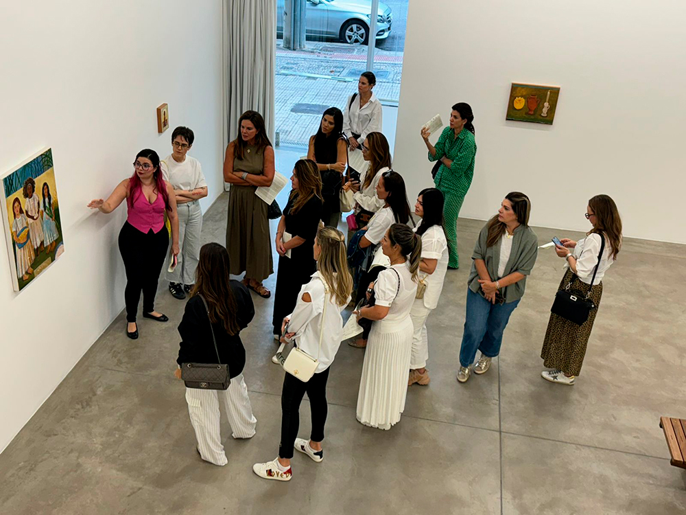 Uma mulher de cabelo rosa está guiando uma visita em uma galeria de arte. Ela veste uma blusa rosa vibrante sem mangas, calça preta e sapatilha preta. Está de lado, fazendo um gesto com a mão, enquanto fala para um grupo de pessoas que a observa. Ao fundo, há três pinturas nas paredes brancas: uma ao lado dela com três mulheres representadas, uma pintura pequena ao fundo, e outra maior do lado direito, que mostra uma composição com frutas. A galeria é bem iluminada, com piso de cimento queimado e uma grande vitrine de vidro ao fundo, por onde dá para ver a rua. As pessoas estão em pé, prestando atenção, muitas segurando papéis, bolsas ou celulares. A maioria veste roupas claras, com destaque para branco e bege.
