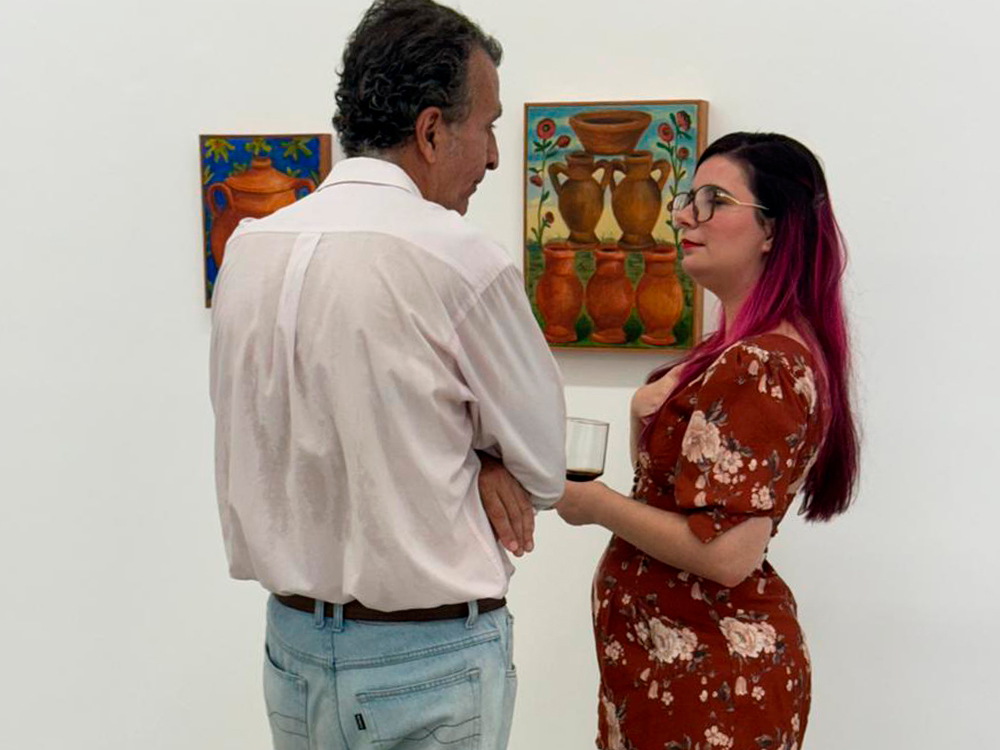 Em uma galeria de arte, uma mulher de cabelos longos e rosa, usando um macaquinho marrom com estampa floral clara, conversa com um homem de cabelos grisalhos e camisa social clara. Eles estão de perfil, voltados um para o outro, e a mulher segura um copo de Coca-Cola. Ao fundo, duas pinturas coloridas estão expostas na parede branca, ambas representando jarros em tons terrosos com elementos decorativos ao redor.