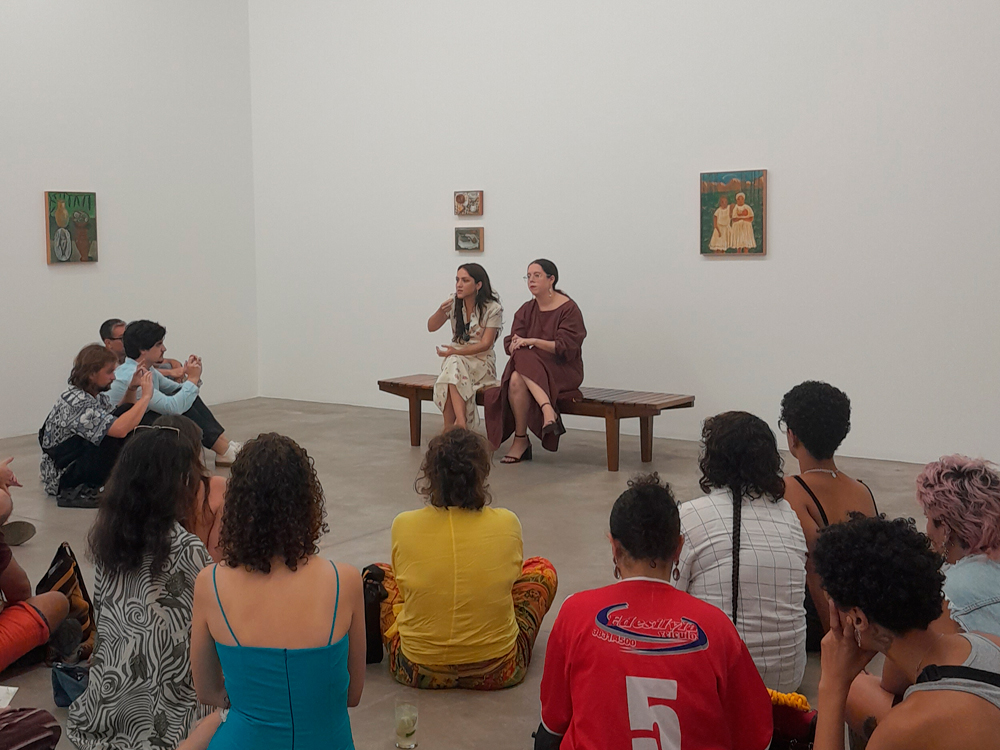 Dentro de uma galeria de arte, duas mulheres estão sentadas em um banco de madeira, conduzindo uma conversa ou roda de conversa. A mulher à esquerda usa um vestido claro estampado, enquanto a da direita, Ayla de Oliveira, veste um vestido marrom. Elas estão de frente para o público, composto por pessoas sentadas no chão, ouvindo atentamente. Ao fundo, em paredes brancas, estão expostas três obras de arte, com composições que incluem jarros, figuras humanas e elementos da natureza.