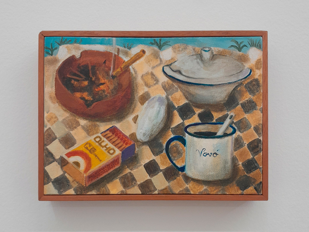 Pintura de Ayla de Oliveira representando uma mesa com toalha quadriculada marrom e bege. Sobre a mesa há uma caneca esmaltada branca com borda azul, escrita ‘Vovó’, com uma colher dentro; uma caixa de fósforos da marca Olho; um cinzeiro marrom com bitucas de cigarro; uma marmita de louça com tampa e um pão francês. Ao fundo, detalhes de grama e céu azul.