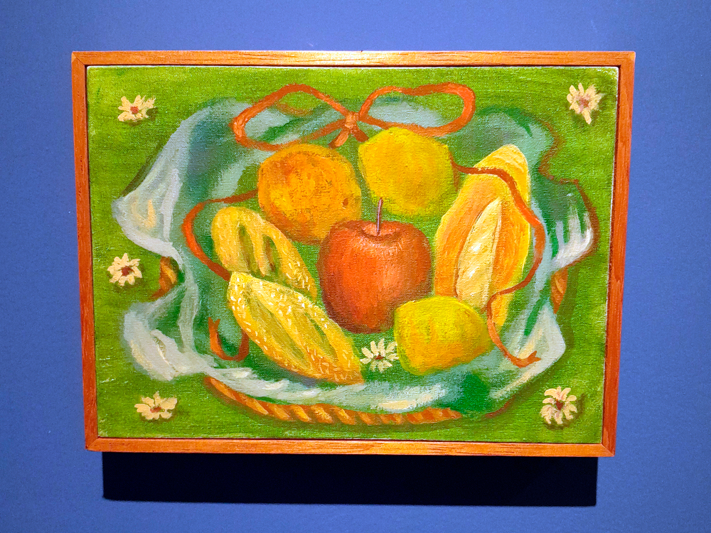 Pintura de uma cesta com frutas sobre um pano azul claro e verde, enfeitado com um laço laranja. Na cesta, há uma maçã vermelha, uma laranja, dois limões, uma banana descascada e uma fruta cortada ao meio, possivelmente um mamão. O fundo é verde com pequenas flores brancas.