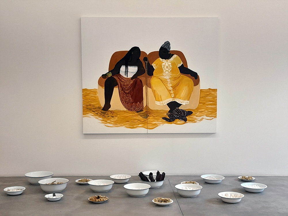 Na parede, uma pintura retratando uma mulher jovem sentada em um sofá ao lado de uma idosa, as duas com os pés em um rio amarelo. Ao pé da mulher há peixes e ao pé da idosa uma tartaruga. Em frente à pintura, no chão, uma instalação com elementos da umbanda , bacias e outros recipientes com miçangas, sal grosso, carvão e água.