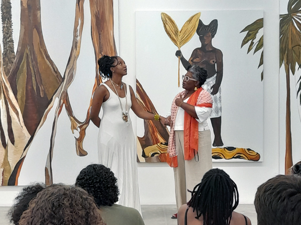 Ao fundo, o tríptico da floresta na parede. Em frente a ele estão duas mulheres negras que vestem roupas claras, sendo elas Hariel, a artista, e Ana Paula, autora do texto curatorial. Em frente a elas, de costas para a câmera, são visível na parte inferior da foto cabeças de pessoas que escutam sua fala