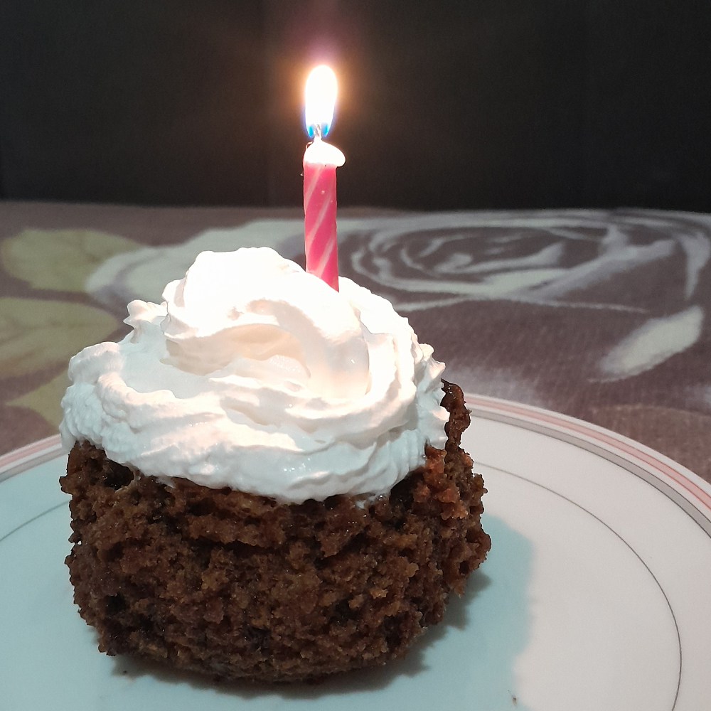 Fotografia em close de um pequeno bolo de chocolate com textura úmida e rústica. No topo, há uma generosa porção de chantili branco, em formato espiralado. Uma vela rosa com listras brancas está acesa bem no centro do chantili. A chama da vela brilha com um leve reflexo azul na base. O bolo está sobre um prato branco com borda fina em rosa e cinza.