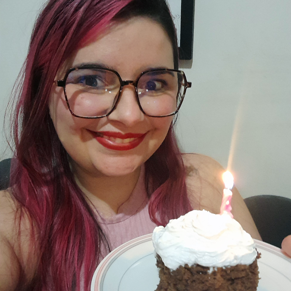 Mulher de cabelos longos tingidos de rosa escuro e pele clara sorri para a câmera segurando um prato com um pequeno bolo de aniversário do blog, sabor chocolate decorado com chantili branco e uma vela rosa acesa. Ela usa óculos de armação grande e maquiagem com batom vermelho vibrante. Veste uma blusa sem mangas em tom rosado. O ambiente tem paredes claras e iluminação suave, e a expressão da mulher transmite alegria e celebração.