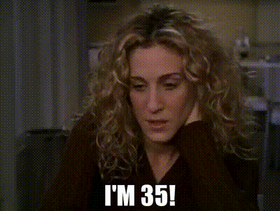 Cena da série Sex and the City. A personagem Carrie Bradshaw, uma mulher branca de cabelos loiros cacheados e soltos, está sentada, apoiando o rosto na mão, com expressão preocupada e um pouco angustiada, dizendo TENHO TRINTA E CINCO ANOS! Na legenda, em letras maiúsculas, aparece a frase descrita.