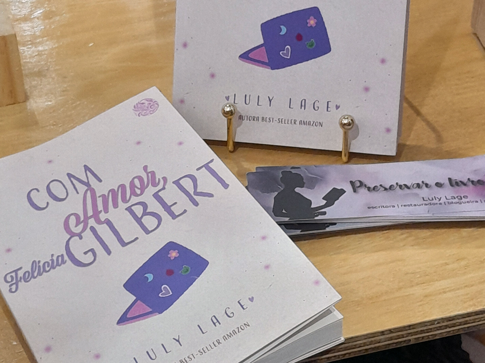 Fotografia de uma mesa de madeira exibindo materiais relacionados ao livro Com Amor, Felícia Gilbert, de Luly Lage. Em primeiro plano, vários exemplares do livro estão empilhados, com a capa em tons de lilás e rosa trazendo o título em destaque e a ilustração de um notebook roxo decorado com pequenos adesivos. Ao fundo, um expositor vertical repete a arte da capa e identifica a autora como best-seller da Amazon. Ao lado dos livros, há marcadores de página personalizados com a frase “Preservar livros” e o nome da autora.