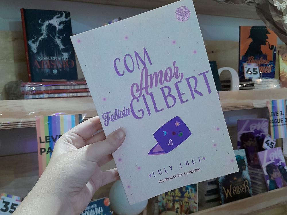 Fotografia em primeiro plano de uma mão segurando o livro. Ao fundo, desfocados, vê-se prateleiras de madeira com outros livros expostos, incluindo exemplares de Love, Sussurros do Abismo e Walker, além de cartazes promocionais com faixas coloridas e a frase Leve 3, Pague 2.