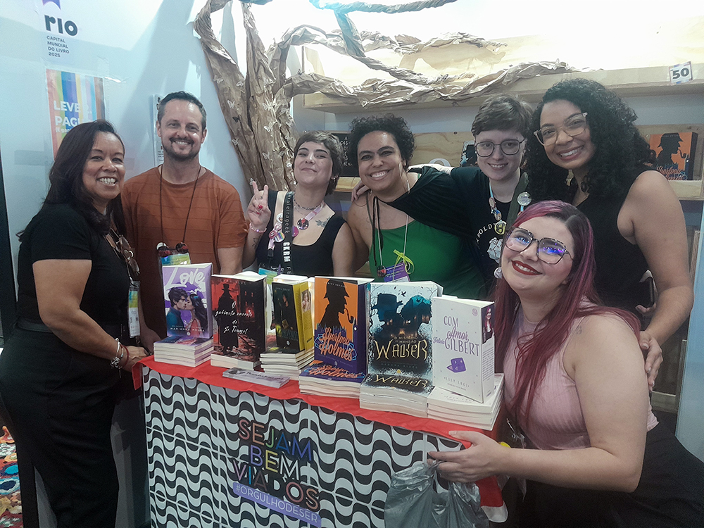 Fotografia de um grupo de sete pessoas sorrindo atrás de uma mesa repleta de livros. Em primeiro plano, sobre a mesa, há pilhas de exemplares organizados de forma destacada A mesa está coberta por uma toalha com estampa ondulada em preto e branco, com as palavras SEJAM BEM-VIADOS e a hashtag #ORGULHODESER. Ao fundo, há uma árvore cenográfica feita de papel e prateleiras de madeira com mais livros. Todas as pessoas na foto estão sorrindo; algumas usam crachás e acessórios coloridos. Luly Lage aparece à frente, agachada ao lado direito da imagem, com cabelos e blusa rosa, sorrindo diretamente para a câmera.