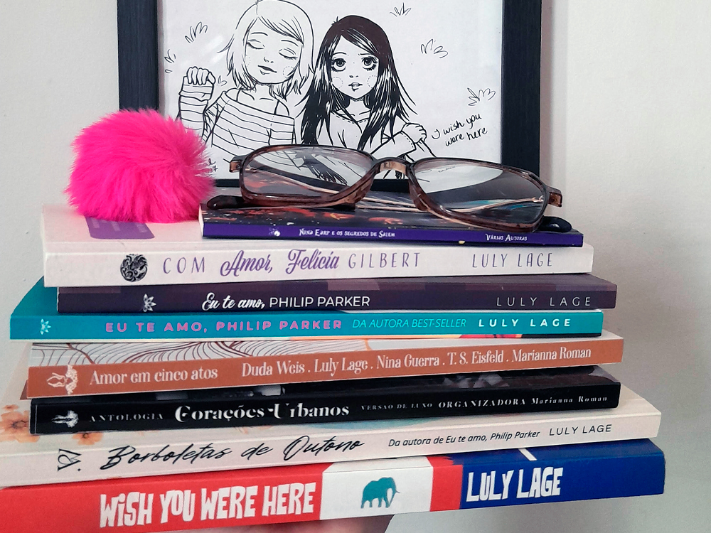 Uma pilha de livros está organizada horizontalmente sobre uma superfície clara. Todos os livros foram escritos por Luly Lage e representam suas publicações ao longo dos anos. No topo da pilha, há um chaveiro de pompom rosa-choque e um par de óculos de grau. Ao fundo, apoiada na parede, há uma ilustração em preto e branco de duas meninas desenhadas no estilo mangá. Uma delas segura um cartaz com a frase “I wish you were here”.