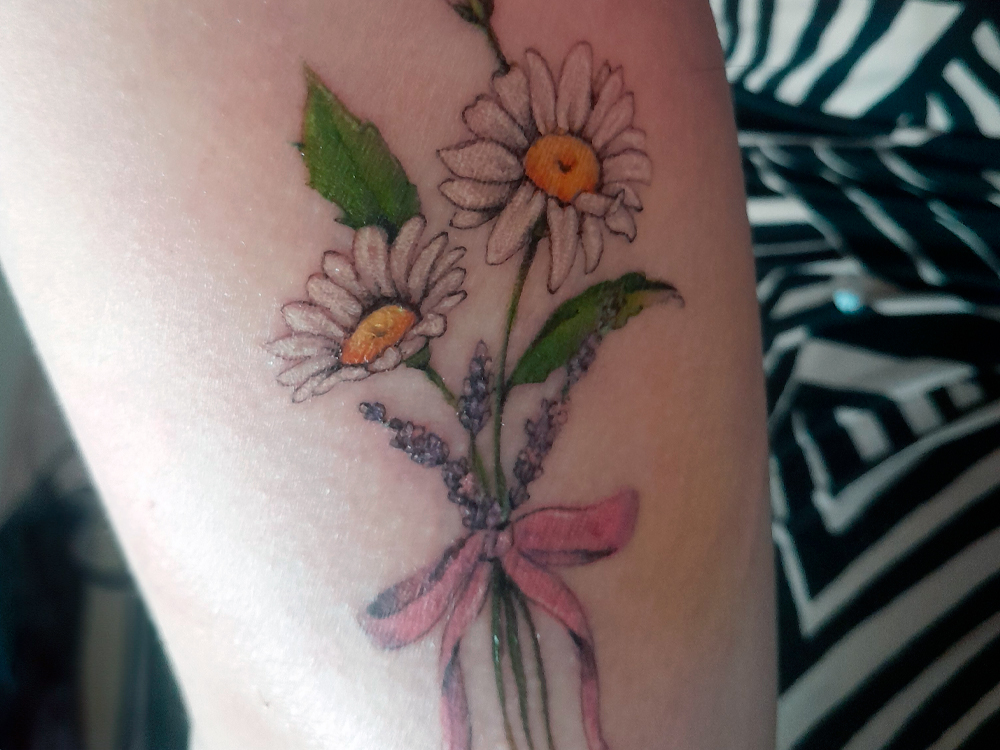 Close da tatuagem, focando nos detalhes do desenhode margaridas com miolo amarelo e pétalas brancas, acompanhadas por pequenas flores lilás (lavandas) e um laço cor-de-rosa amarrando os caules. A imagem mostra que a tatuagem está na fase de cicatrização, com uma leve casquinha visível sobre as flores. O fundo da foto é composto por um tecido de estampa listrada em preto e branco, parte da roupa da pessoa.