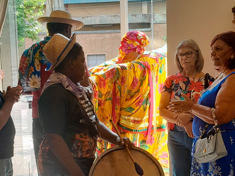 Cena animada de um grupo reunido durante a exposição. Um percussionista toca tambor, enquanto outras pessoas, vestindo roupas coloridas e estampadas, interagem próximas à entrada iluminada pela luz natural. Ao fundo, uma figura usa um traje vibrante de boi amarelo com flores, trazendo um ar festivo ao momento.