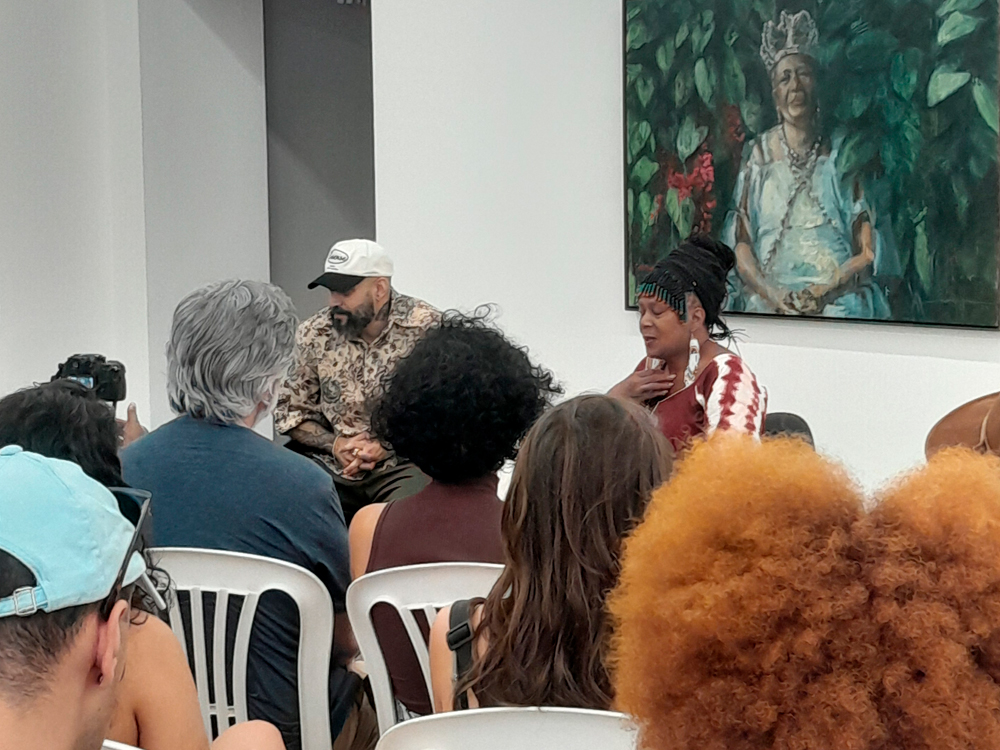Registro de uma roda de conversa durante a exposição. Diego Mouro, um homem de boné claro e camisa estampada e uma mulher com tranças e vestido listrado participam do bate-papo, sentados à frente do público. Ao fundo, um quadro retrata uma figura feminina coroada, cercada por folhagens.