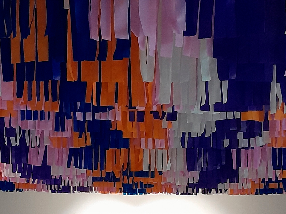 Detalhe do teto da exposição com fitas de papel coloridas penduradas, criando um efeito vibrante e festivo. As tiras aparecem em tons de roxo, laranja, rosa e branco, formando uma composição cheia de movimento e textura.