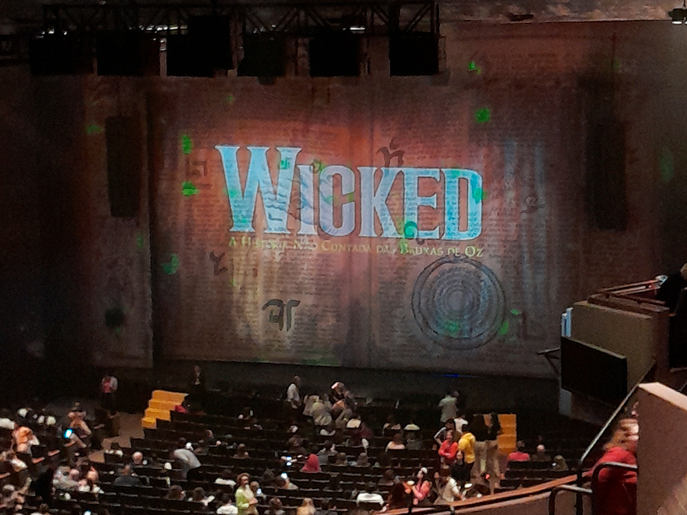 Foto tirada do balcão superior do Teatro Renault, em São Paulo, mostrando o palco antes do início da apresentação do musical Wicked. A cortina de fundo traz o nome do espetáculo em destaque: WICKED – A História Não Contada das Bruxas de Oz, projetado em tons esverdeados sobre um pano de fundo com textura de livro antigo e símbolos místicos. A plateia ainda está se acomodando, com pessoas caminhando entre as fileiras.