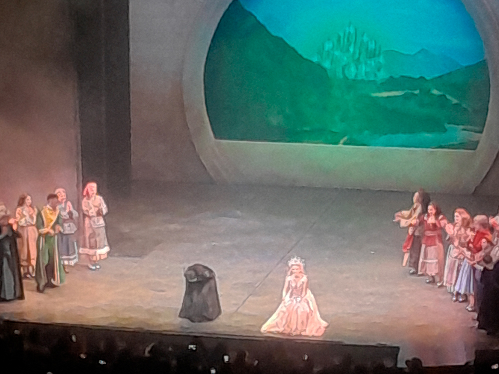 Momento dos agradecimentos finais do musical Wicked, no palco do Teatro Renault. Glinda, a Boa, está ajoelhada no centro do palco, com seu vestido rosa claro e tiara brilhante, sorrindo para o público. Ao seu lado, uma figura encapuzada em preto – representando Elphaba, a Bruxa Má do Oeste – faz uma reverência. Ao redor delas, o restante do elenco forma um semicírculo, aplaudindo e sorrindo, vestindo os figurinos dos cidadãos de Oz. Ao fundo, uma paisagem com o Castelo de Oz completa o cenário. Na parte inferior da imagem, vê-se a orquestra e a plateia aplaudindo de pé, com algumas pessoas levantando os celulares para registrar o momento de encerramento.