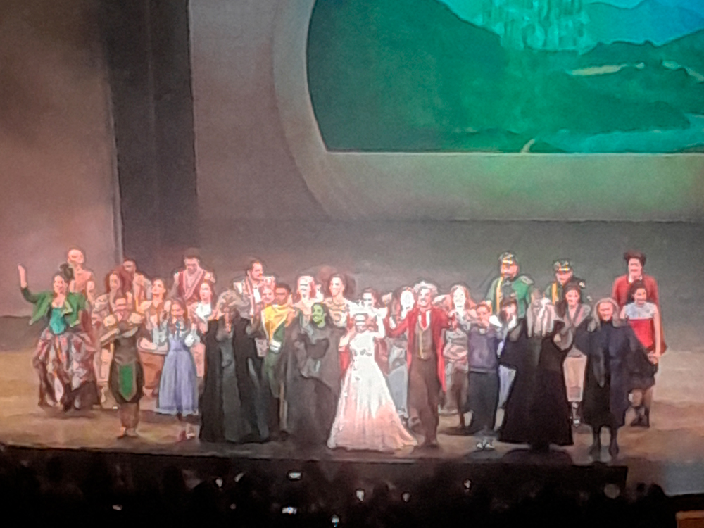 Elenco completo de Wicked reunido no palco durante os aplausos finais no Teatro Renault, em São Paulo. Todos estão alinhados em frente ao cenário que exibe uma paisagem com o Castelo de Oz ao fundo. No centro da imagem estão Elphaba, com a pele verde característica e vestido escuro, e Glinda, com um vestido claro e brilhante, ambas sorrindo e aplaudindo junto ao grupo. Atrás e ao redor delas, os demais integrantes do elenco — usando figurinos coloridos inspirados nos cidadãos de Oz — também aplaudem o público, acenam e sorriem.